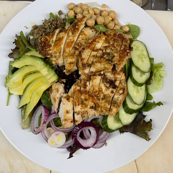 Grilled chicken salad with avocado, cucumbers, red onion, chickpeas, and quinoa on a white plate.