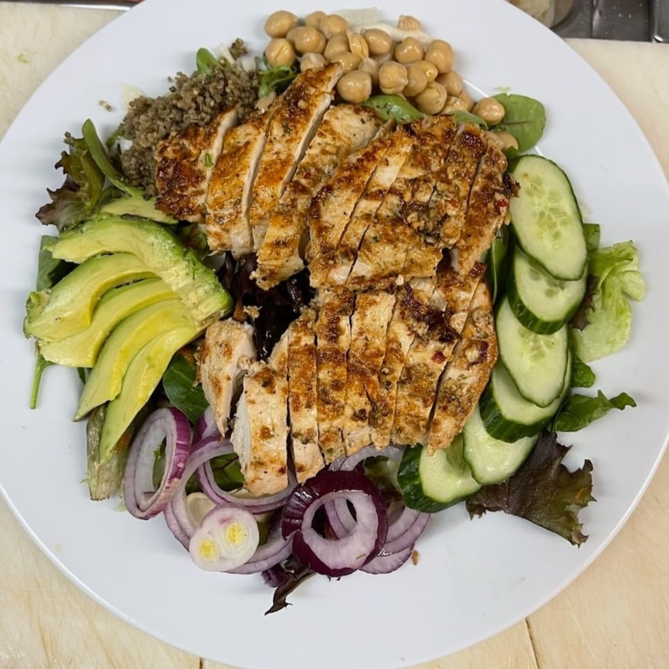 Grilled chicken salad with avocado, cucumbers, red onions, chickpeas, and quinoa on a white plate.