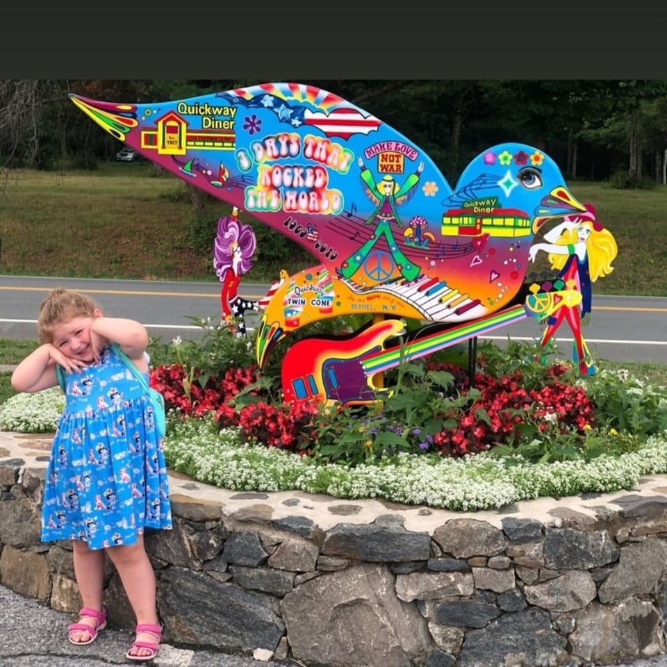 A young girl poses beside a colorful, bird-shaped Quickway Diner sign with Woodstock-themed art.