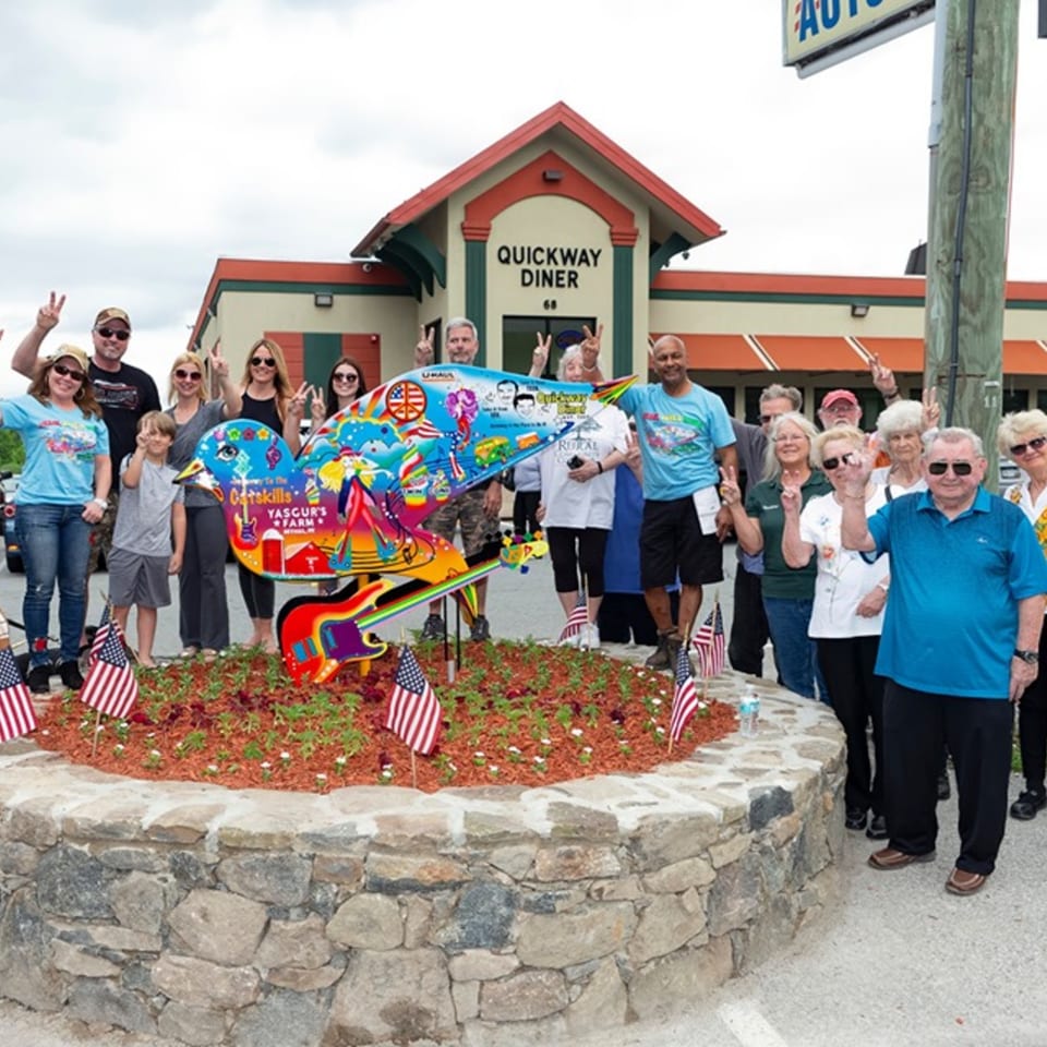People giving peace signs around a colorful bird sculpture with guitars in front of Quickway Diner.