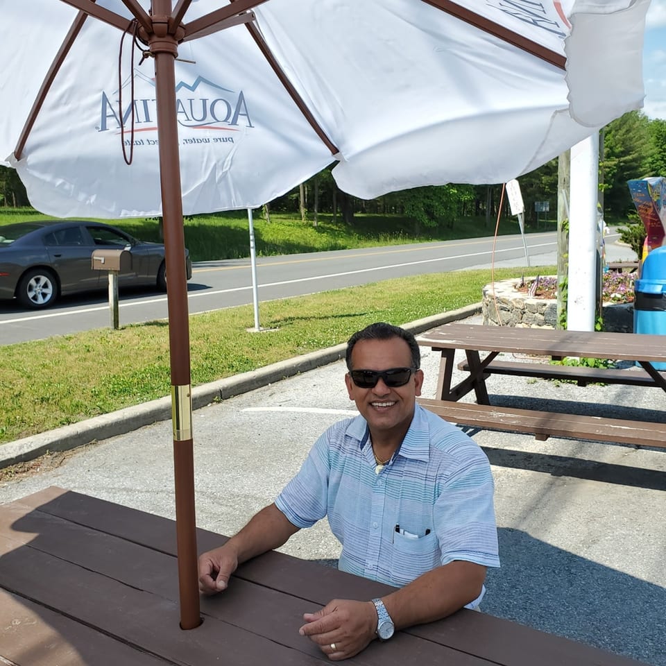 Man in sunglasses smiling at a Quickway Diner outdoor picnic table under an umbrella.
